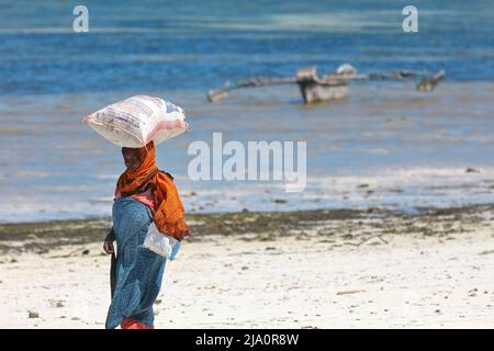 Une femme portant un sac d'algues sur sa tête à la plage de Jambiani avec un bateau traditionnel 'Ngalawa' en arrière-plan, Zanzibar, Tanzanie, Afrique. Banque D'Images