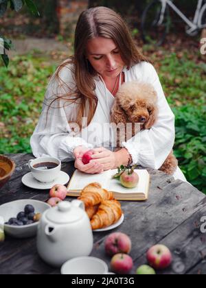 femme de race blanche d'âge moyen en robe blanche prenant le café du matin dans le verger de pomme assis à une table en bois avec son animal de compagnie Banque D'Images