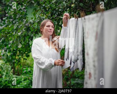 femme caucasienne d'âge moyen en robe blanche faisant des devoirs, pendant des vêtements sur la corde à linge dans la rue dans la cour de la maison de village, jour de blanchisserie Banque D'Images