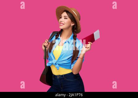 Femme caucasienne souriante étudiante avec passeport et billets d'avion. Fille en vêtements décontractés et chapeau de paille. Femme voyageur sur fond rose isolé. Banque D'Images