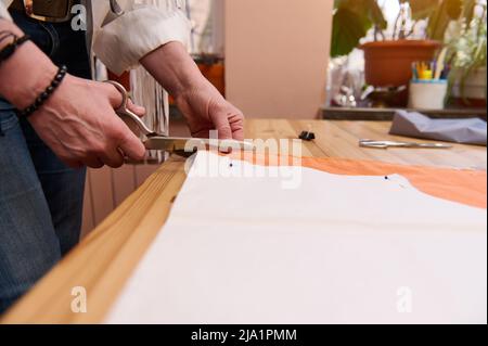 Gros plan des mains de styliste de mode utilisant des ciseaux de coupe le motif de couture dans le tissu de satin de couleur orange dans l'atelier de tailleur. Couturière à partir d'une ne Banque D'Images