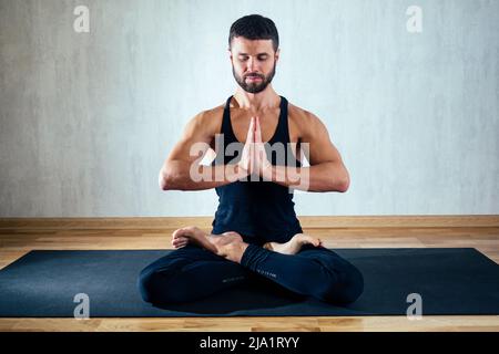un homme en vêtements de sport sombres pratiquant le yoga dans une position de lotus sur fond gris. asana sur le sol sur des tapis de yoga. le concept de concentration Banque D'Images