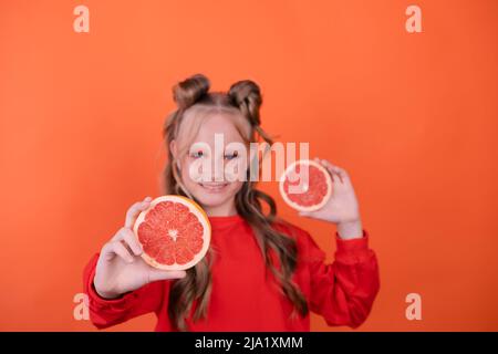 Jolie petite fille à l'orange avec un pamplemousse isolé sur fond orange. Agrumes tropicaux tranches de pamplemousse. Orange élégant maquillage. Adolescent Banque D'Images