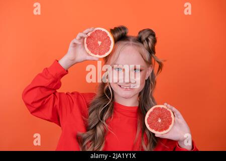 Jolie petite fille à l'orange avec un pamplemousse isolé sur fond orange. Agrumes tropicaux tranches de pamplemousse. Orange élégant maquillage. Adolescent Banque D'Images