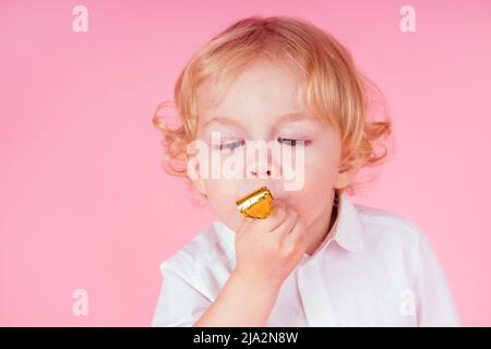 petit garçon blond avec boucles coiffure 4-5 ans dans le studio sur un fond rose bourdonneurs sifflent un coup de sifflet un anniversaire fête célèbre Banque D'Images