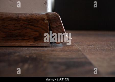 Gros plan du coin de la plinthe endommagé avec de la poussière et des fils de toile d'araignée sur un plancher en bois, avec un mur rayé blanc et un arrière-plan sombre Banque D'Images