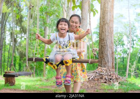 La jeune mère balance pour sa fille dans le parc. Petite fille souriante assise sur balançoire sous l'arbre avec mère dans le parc d'été. Bonne famille s'amuser Banque D'Images