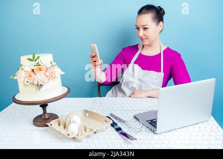 la femme au foyer (pâtisserie-cuisinière) prend des photos de gâteau d'anniversaire blanc crémeux à deux niveaux (mariage) avec des fleurs fraîches au téléphone et un Banque D'Images