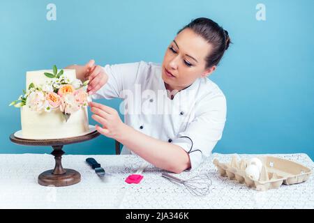 la confiseur (pâtisserie-pâtisserie) décorez l'appétissant gâteau de mariage blanc crémeux à deux niveaux avec des fleurs fraîches sur une table avec une nappe en dentelle Banque D'Images