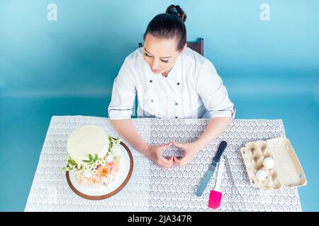 le chef pâtissier affiche le symbole du cœur des doigts à côté du gâteau de mariage (anniversaire) blanc crémeux à deux niveaux avec des fleurs fraîches sur la table Banque D'Images