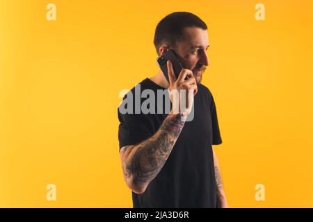Jeune homme parlant au téléphone, bras tatoués, regardant loin studio tourné espace de copie isolé fond jaune . Photo de haute qualité Banque D'Images