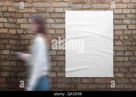 Une fille passe devant une affiche blanche sur un mur de rue en brique. Affiche blanche vierge avec texture froquée pour la présentation du design Banque D'Images