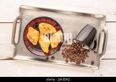 Quatre morceaux de baklava au miel sucré avec une soucoupe en céramique, une tasse, des grains de café et un plateau en métal sur une table en bois, vue rapprochée du dessus. Banque D'Images