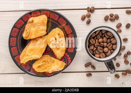 Quelques morceaux de baklava au miel sucré sur une soucoupe en céramique, avec des grains de café dans une tasse sur une table en bois, vue rapprochée, vue du dessus. Banque D'Images