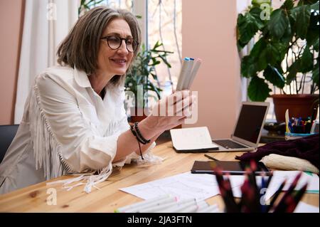 Femme européenne mûre et confiante, designer de mode tenant des marqueurs d'aquarelle tout en étant assise sur un bureau en bois et en créant de nouvelles esquisses de vêtements à la mode Banque D'Images