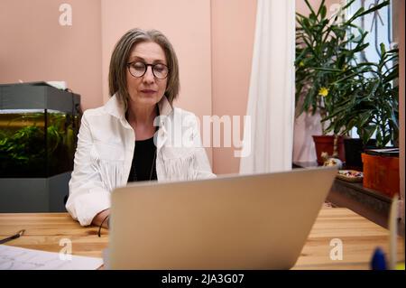 Femme européenne âgée designer de mode, couturière, travail sur mesure sur ordinateur portable, recevoir des commandes en ligne assis sur un bureau en bois dans un atelier de design de mode Banque D'Images