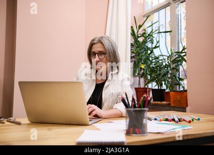 Femme européenne mûre et confiante, couturière à l'aide d'un ordinateur portable et en concevant de nouveaux vêtements pour une nouvelle collection, assise sur un bureau en bois dans un Banque D'Images