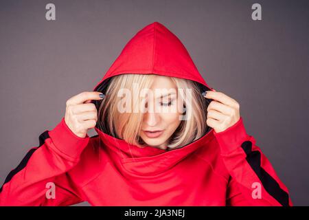 portrait d'une belle femme blonde peau parfaite et maquillage mettre sur la capuche sweat-shirt rouge survêtement sport costume hip hop danseuse en studio sur noir Banque D'Images