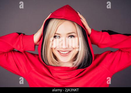 portrait d'une belle femme blonde peau parfaite et maquillage mettre sur la capuche sweat-shirt rouge survêtement sport costume hip hop danseuse en studio sur noir Banque D'Images