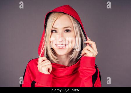 portrait d'une belle femme blonde peau parfaite et maquillage mettre sur la capuche sweat-shirt rouge survêtement sport costume hip hop danseuse en studio sur noir Banque D'Images