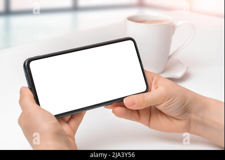 maquette de la position horizontale du téléphone portable. Image de maquette d'une femme tenant un téléphone portable noir avec un écran vierge et une tasse de café sur une table. Gros plan de la femme ha Banque D'Images