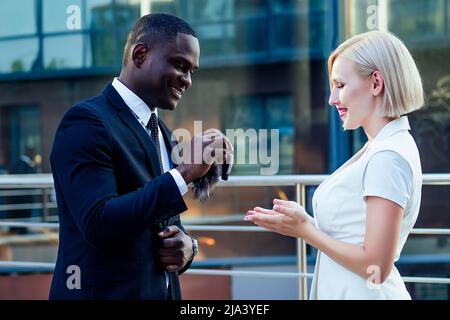 Beau homme d'affaires afro dans un costume noir élégant donne des clés de voiture à la belle blonde femme d'affaires sur fond de rue gratte-ciel de bureau. Louer et Banque D'Images