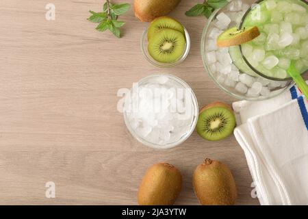 Arrière-plan avec détail de la boisson sans alcool kiwi avec beaucoup de glace sur un banc de cuisine en bois avec des fruits et un bol avec de la glace autour d'elle. Vue de dessus. Horizontale c Banque D'Images