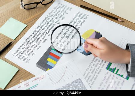 vue rognée du directeur financier tenant la loupe au-dessus des tableaux sur le bureau Banque D'Images