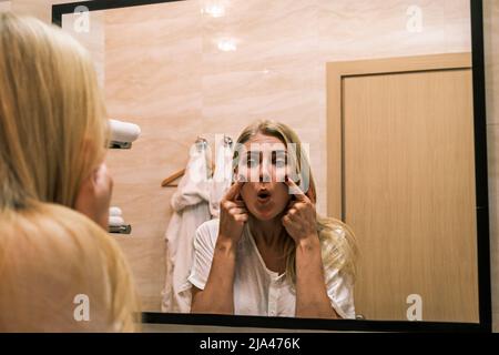 Deuxième menton, obésité. Portrait d'une femme de 30 ans soulevant ses joues en utilisant ses doigts tout en regardant le miroir dans la salle de bains. Vieillissement et apparence Banque D'Images