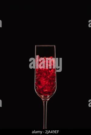 Flûte à champagne remplie de cristaux en forme de coeur rouge transparent rétroéclairés et isolés sur fond noir. Concept d'amour et de célébration. Copier l'espace. Banque D'Images