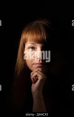 Portrait d'une belle et sérieuse femme d'âge moyen caucasien visage tenant le menton dans l'obscurité d'un côté et éclairé de l'autre côté Banque D'Images