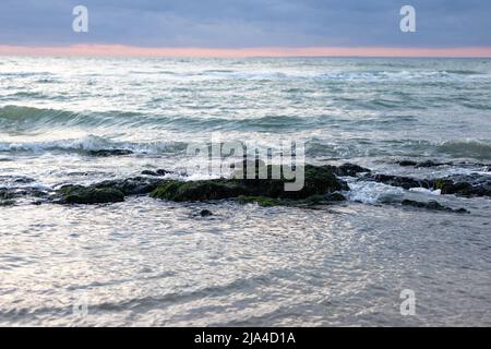 Un beau coucher de soleil rose coloré avec un ciel bleu dramaturge et l'eau de mer éclaboussant légèrement sur les rochers et les pierres. Un paysage magnifique et spectaculaire mais apaisant. Photo de haute qualité Banque D'Images