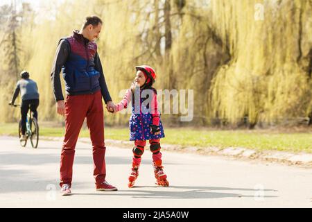 Père enseignant à sa fille de skate sur patins à roulettes. Joyeux enfant dans l'apprentissage du patinage sur casque. Famille passant du temps ensemble. Jour d'été ensoleillé Banque D'Images