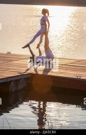 Beau jeune couple faisant un acro yoga sur la jetée contre le coucher du soleil. Homme couché sur la jetée et femme d'équilibrage dans ses pieds. Banque D'Images