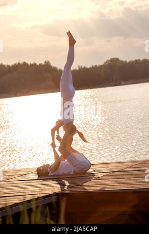 Beau jeune couple faisant un acro yoga sur la jetée contre le coucher du soleil. Homme couché sur la jetée et femme d'équilibrage dans ses pieds. Banque D'Images