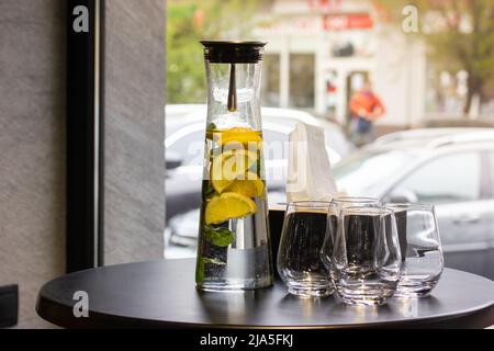 Une carafe en verre transparent, un pichet de limonade avec des citrons, de la menthe à côté des verres sur une table ronde noire dans un café ou un restaurant contre une fenêtre panoramique. A r Banque D'Images