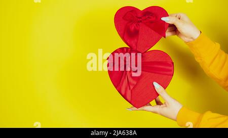 Mains de femme avec des boîtes-cadeaux rouges sous forme de coeur sur fond jaune. Personne méconnaissable tenant des cadeaux romantiques avec un ruban. Concept de présent Banque D'Images