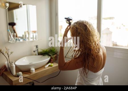 Arrière sur jeune blonde caucasienne femme dans une serviette sèche cheveux avec sèche-cheveux dans la salle de bains. Banque D'Images