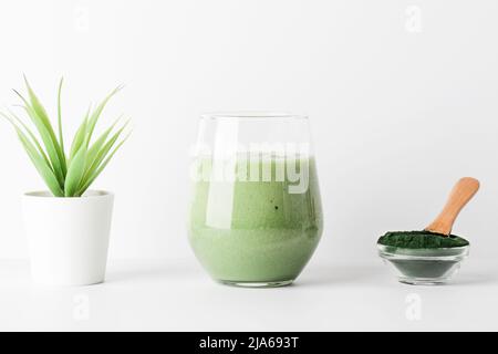 smoothie vert en verre et poudre d'algues spiruline dans un bol avec cuillère, fleur sur fond blanc. concept d'auto-soin et de mode de vie sain. Banque D'Images