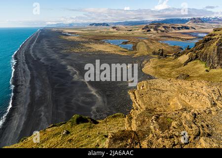 Le promontoire de Dyrhólaey offre ici une vue sur une plage de sable noir volcanique d'un mile de long s'étendant vers l'ouest le long de la côte sud de l'Islande ; des zones humides, un petit lac et des montagnes enneigées se trouvent en arrière-plan. Le Dyrhólaey, haut de 120 mètres, était à l'origine une île volcanique du Pléistocène et tire son nom, signifiant « île de la colline de la porte », de la grande arche rocheuse sur son côté est ; il se trouve non loin du village de Vík í Mýrdal. Banque D'Images