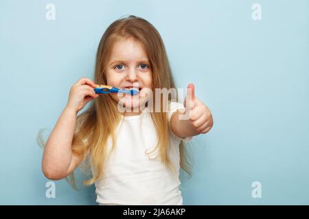 petite fille heureuse se brossant les dents avec une brosse à dents et souriant sur fond bleu. pouce vers le haut Banque D'Images