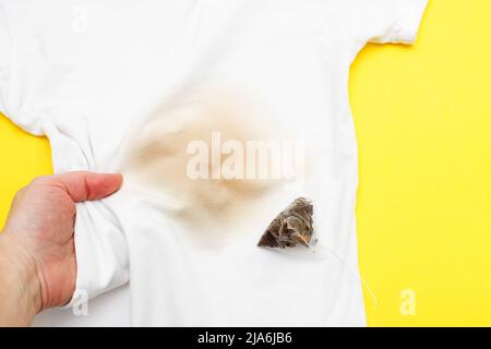 main féminine tenant des vêtements blancs avec une tache de thé sale avec un sac de thé à côté d'elle sur un fond jaune Banque D'Images