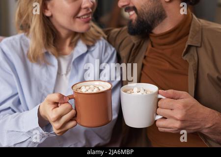 Homme et jeune femme à barbe élégants assis ensemble sur un canapé à la maison, tenant des tasses de café chaud avec guimauve, espace de copie Banque D'Images