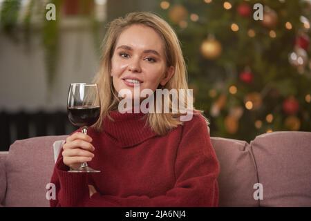 Portrait moyen en gros plan d'une femme attrayante portant un chandail chaud assis sur un canapé dans la salle de séjour décorée pour Noël avec un verre de vin rouge Banque D'Images