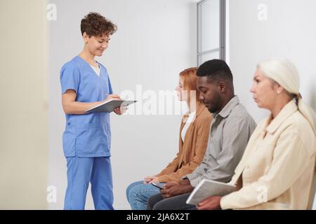 Groupe de trois personnes multiethniques qui attendent une consultation dans un hôpital moderne, un travailleur médical se tenant près de vérifier la présence Banque D'Images