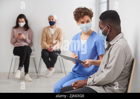Prise de vue horizontale d'une travailleuse médicale méconnue portant un masque de protection assise sur une chaise dans un couloir à côté du patient qui lui parle de test Banque D'Images
