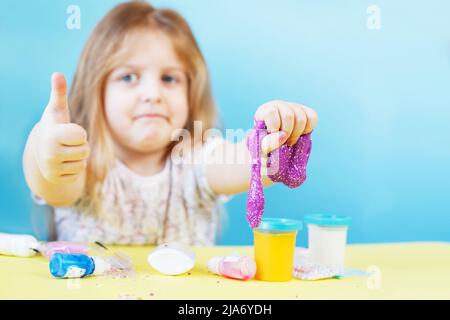 Une fille blonde tient un slime scintillant violet et un signe de make pouce vers le haut isolé sur un fond bleu. Enfant jouant avec un jouet de slime. Faire de la chaux. Copier l'espace. Banque D'Images