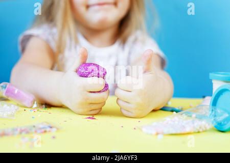 Une fille blonde tient un slime scintillant violet et un signe de make pouce vers le haut isolé sur un fond bleu. Enfant jouant avec un jouet de slime. Faire de la chaux. Copier l'espace. Banque D'Images