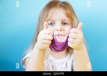 Une fille blonde tient un slime scintillant violet et un signe de make pouce vers le haut isolé sur un fond bleu. Enfant jouant avec un jouet de slime. Faire de la chaux. Copier l'espace. Banque D'Images