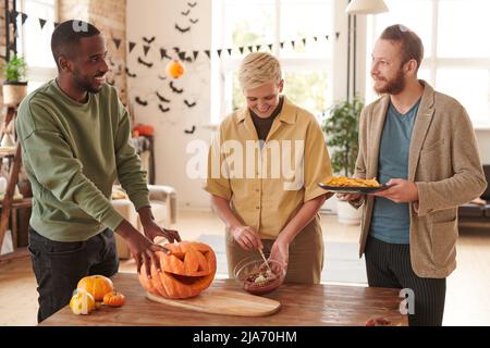 Groupe de jeunes amis multiethniques joyeux debout à table et préparant des en-cas décorés de citrouilles pour la fête d'Halloween Banque D'Images